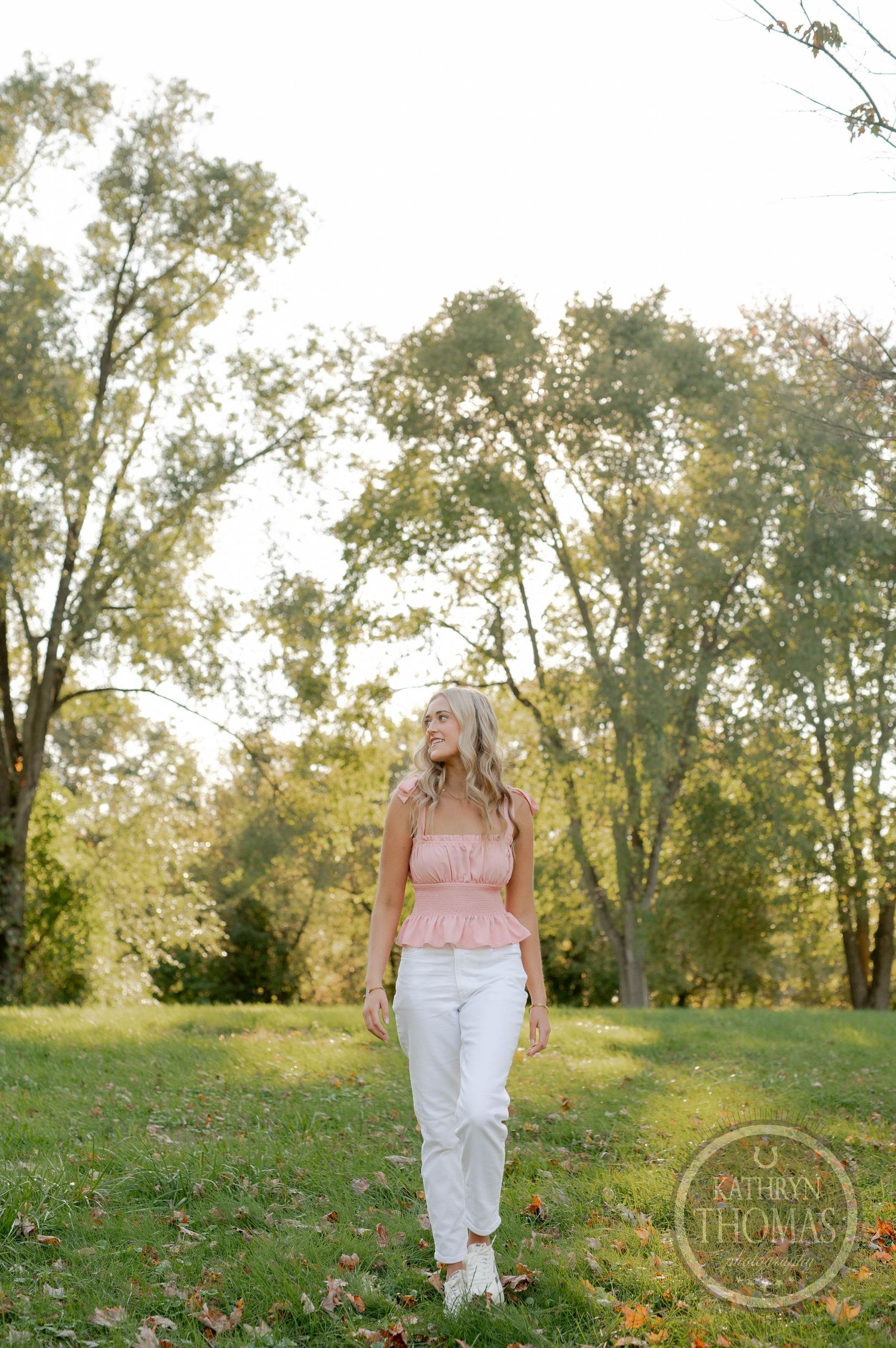Outdoor portrait in a park featuring pink peplum top and white pants against sunlit trees in background.