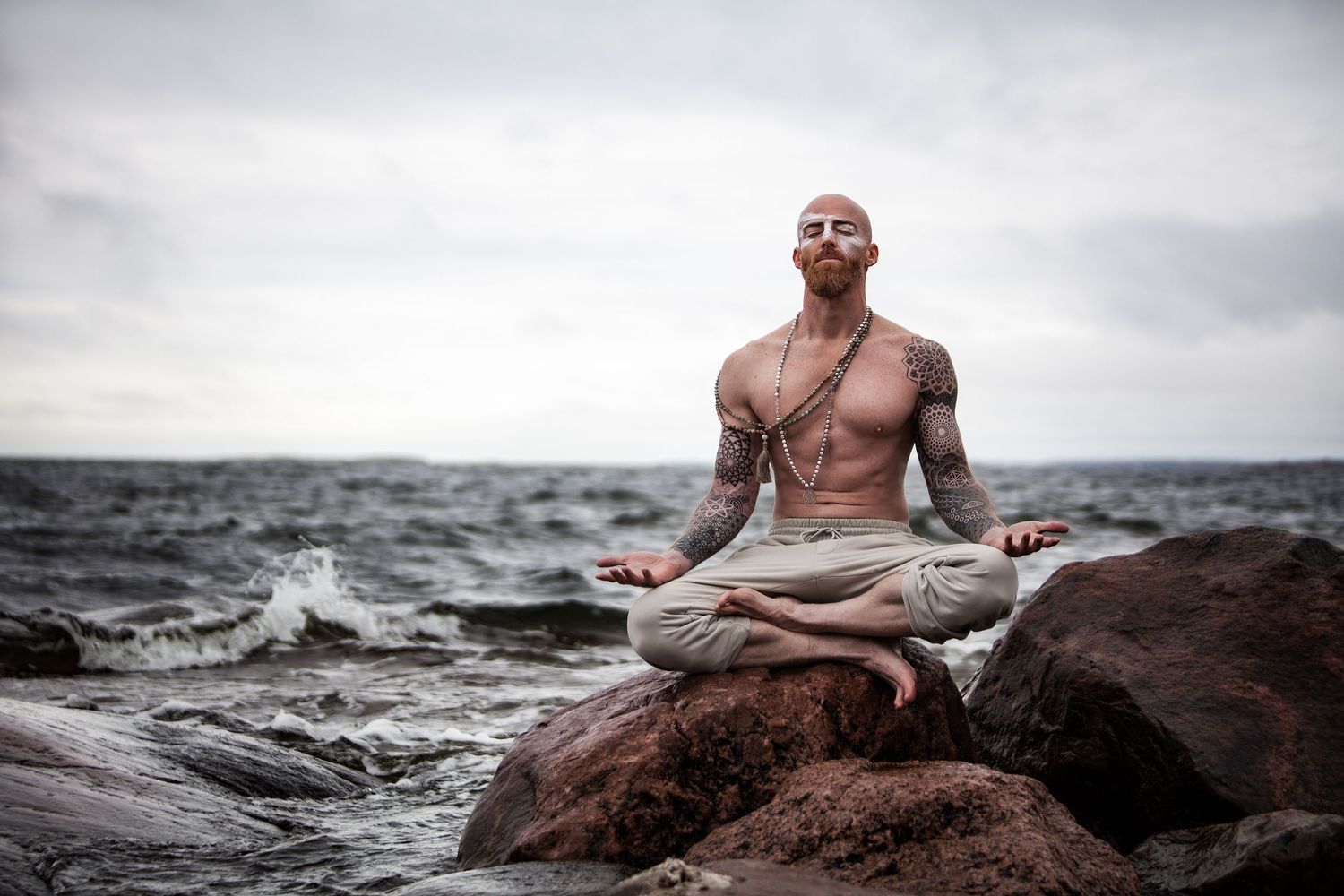 Person meditating in lotus position on rocky shore with dramatic ocean waves in background.