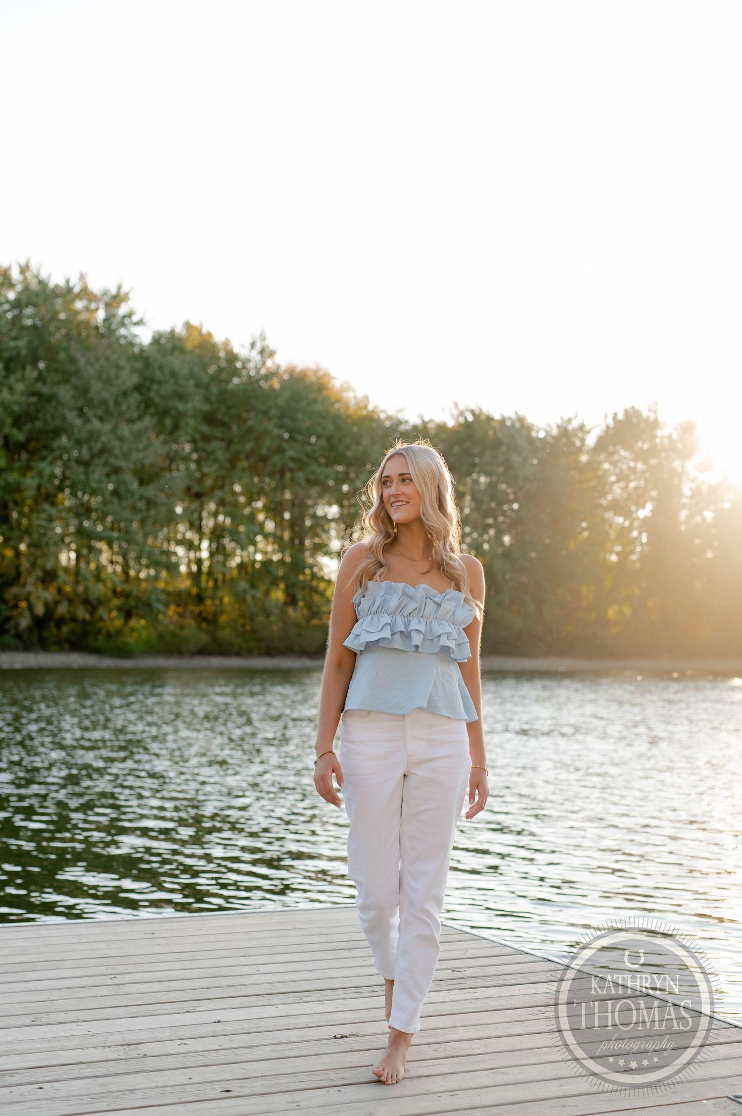 Someone in a light blue top and white pants walking on a wooden dock during golden hour.