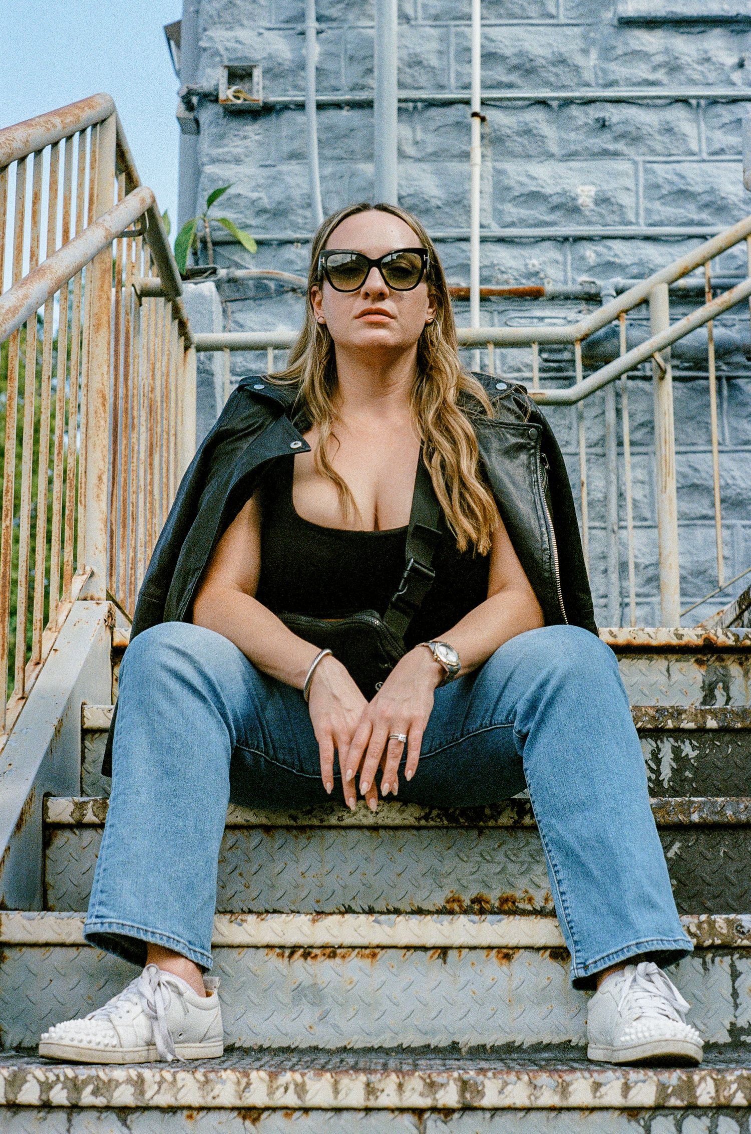 Modern fashion portrait on industrial metal stairs with leather jacket, jeans and sunglasses against stone wall.