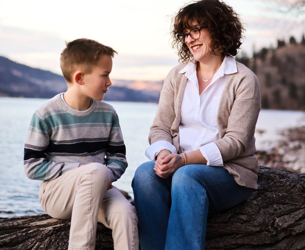 Parent and child share conversation while sitting on rocks by mountain lake at sunset.