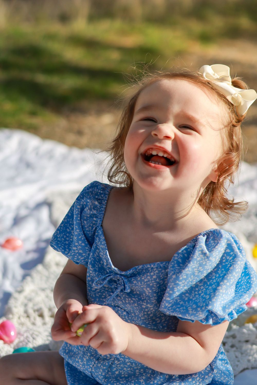 A joyful child wearing a blue polka dot dress and cream bow laughs while sitting outside on a white blanket.
