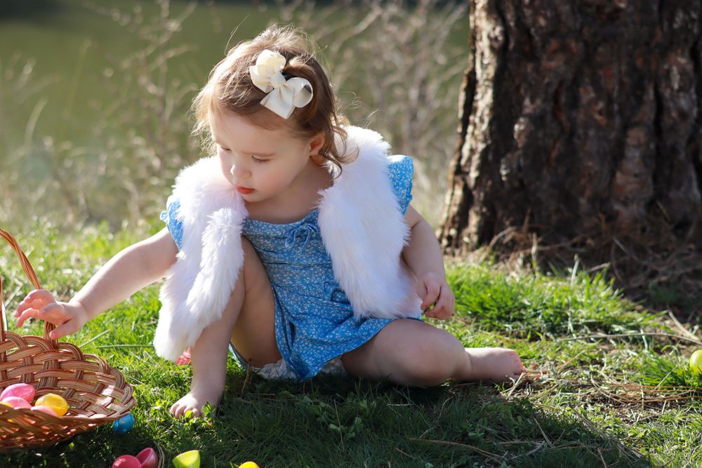 Child in blue dress with white shawl crouches on mossy ground near a tree trunk in dappled sunlight.
