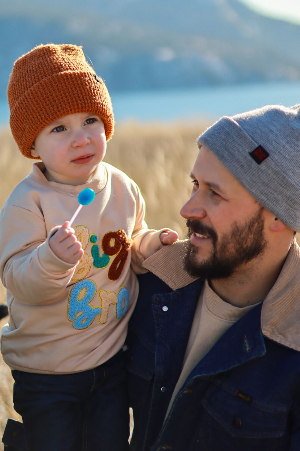 Small child in orange beanie holds a lollipop while being held outdoors on a sunny day.