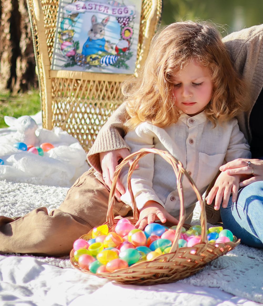 Child sits on blanket with wicker basket filled with colorful plastic Easter eggs during outdoor springtime celebration.