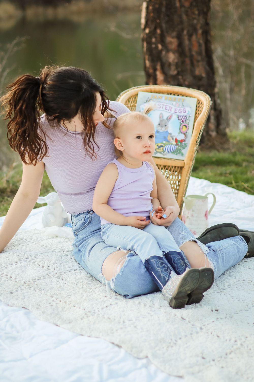 A parent and baby sit on a white blanket outdoors reading a storybook together in matching purple tops and jeans.