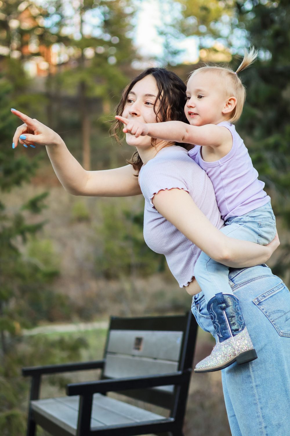 A playful moment outdoors as a toddler rides piggyback and points while being held near a wooden bench in nature.