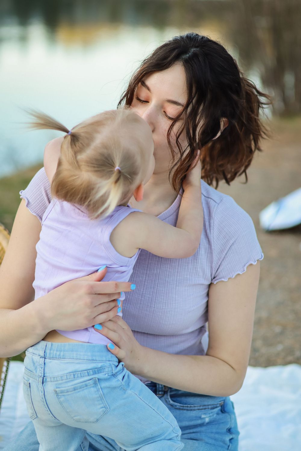 A tender moment captured between parent and child embracing on a blanket outdoors by the water on a peaceful day.