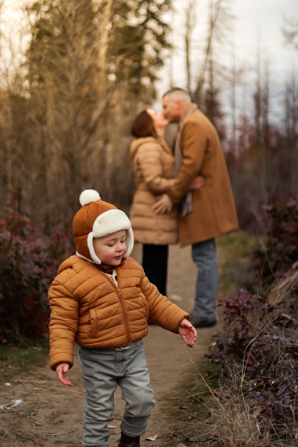 Child in orange puffer jacket walking on path while couple embraces in background.