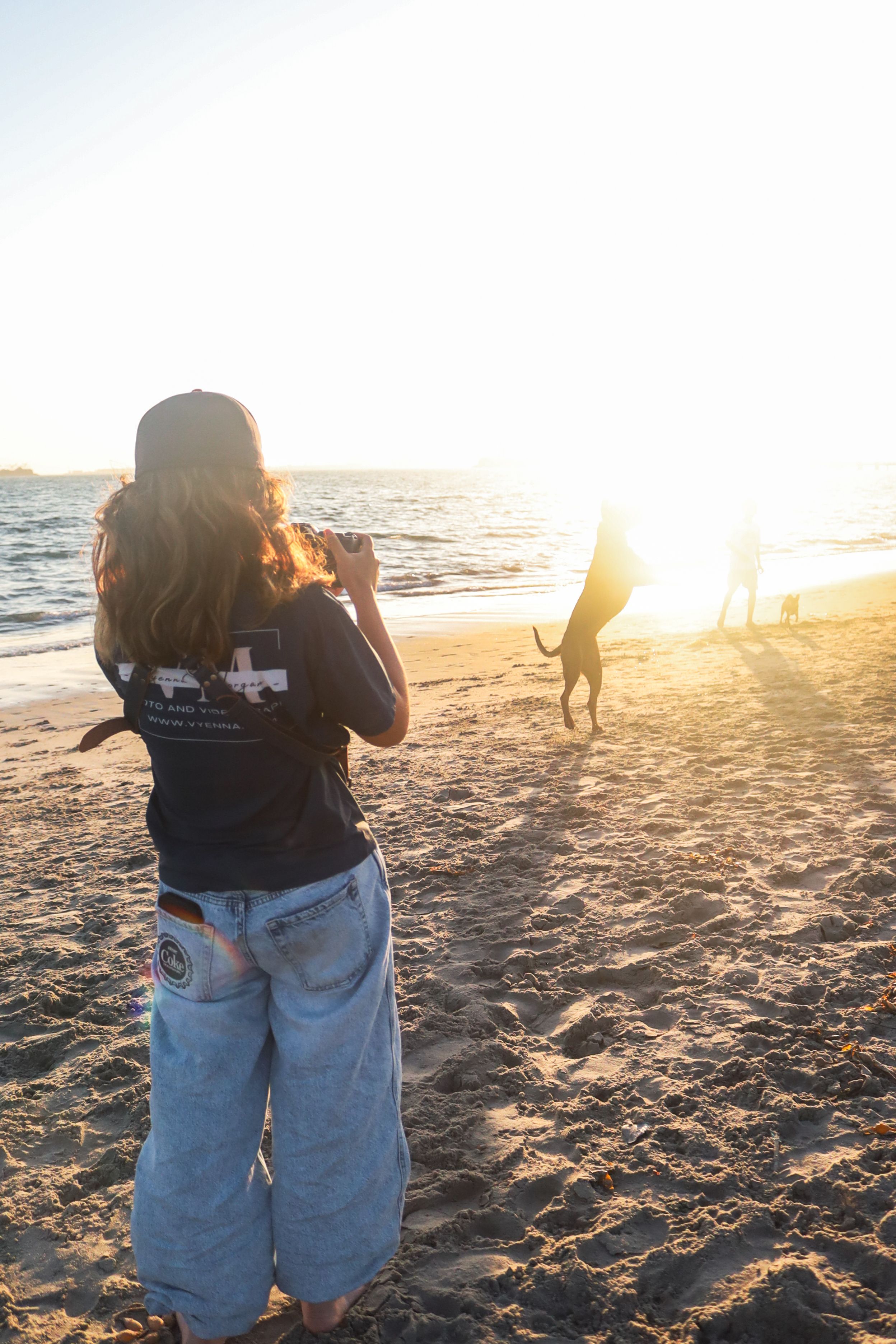 Photographer in baggy jeans captures sunset scenes on a sandy beach with golden light.