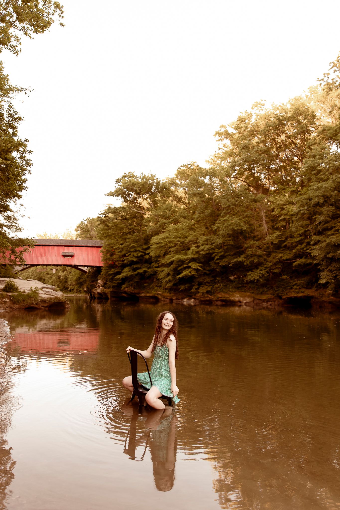 A person in a green dress poses in the water near a red covered bridge surrounded by trees and reflected in the river.
