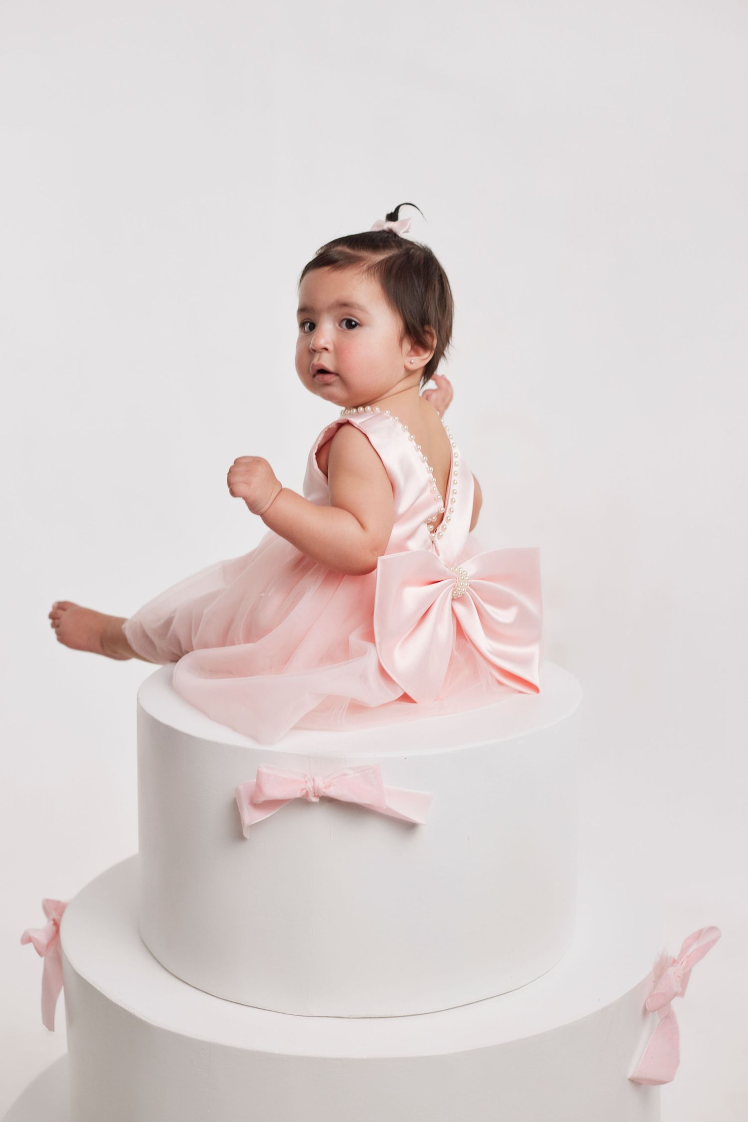 Baby in pink tulle dress with bow sits atop white pedestal.