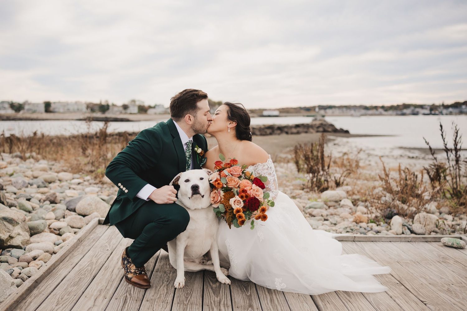 A romantic wedding photo on a wooden deck by the water with a white dog joining the newlyweds.