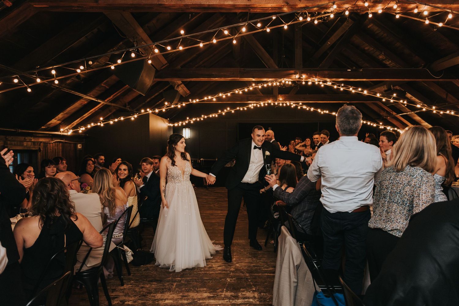 Wedding celebration in a rustic barn venue with string lights overhead as guests dance on the wooden floor.
