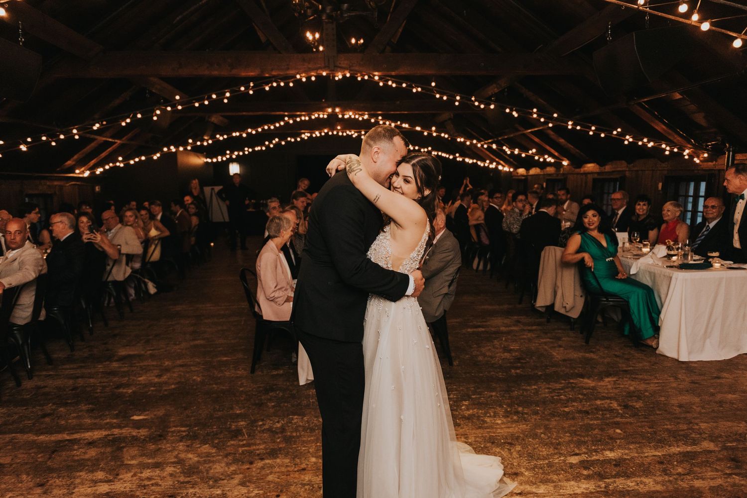 A couple shares their first dance at a rustic barn reception with twinkling lights overhead and guests looking on.