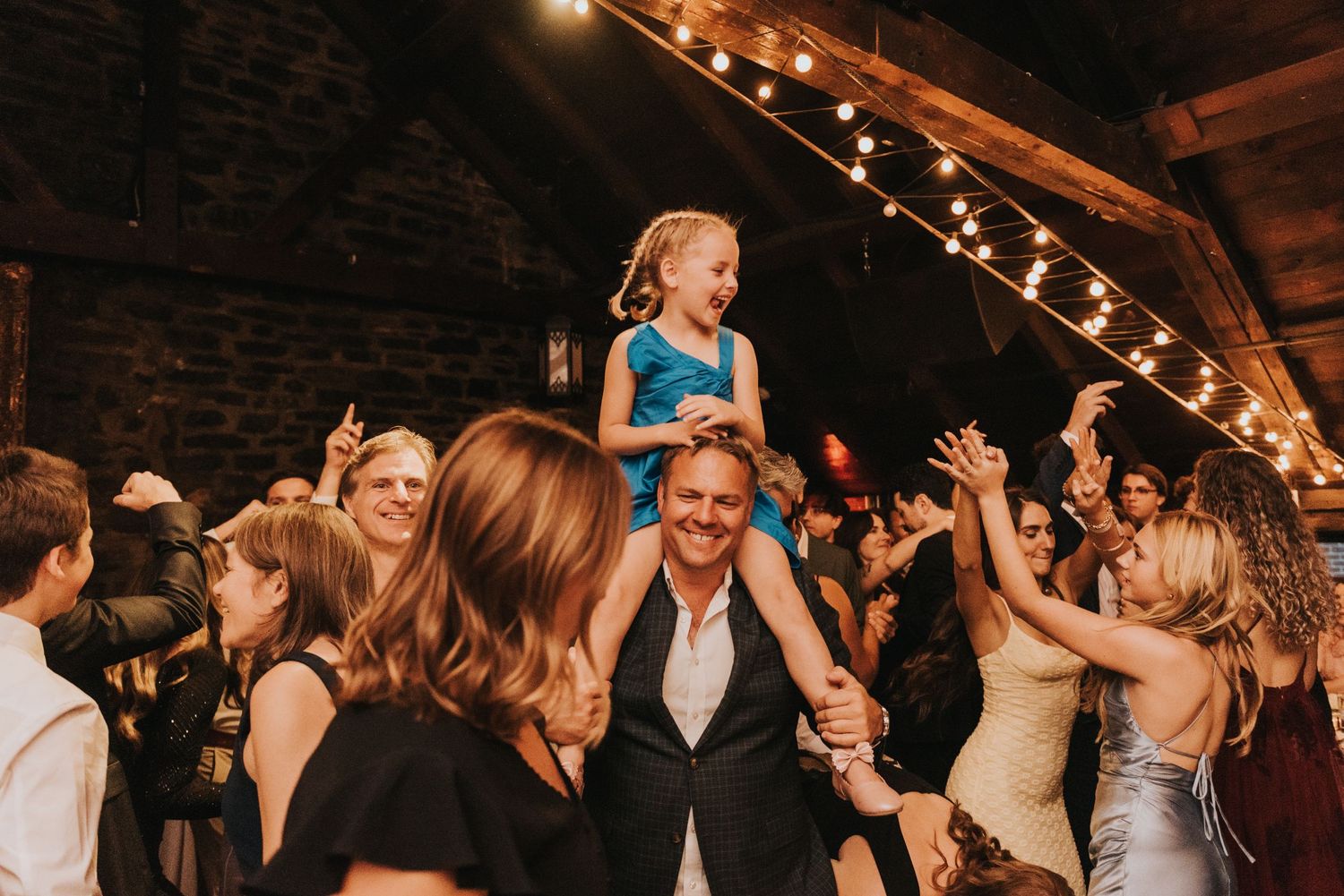 Wedding party celebrates on dance floor as young child sits on adult's shoulders under rustic string lights.
