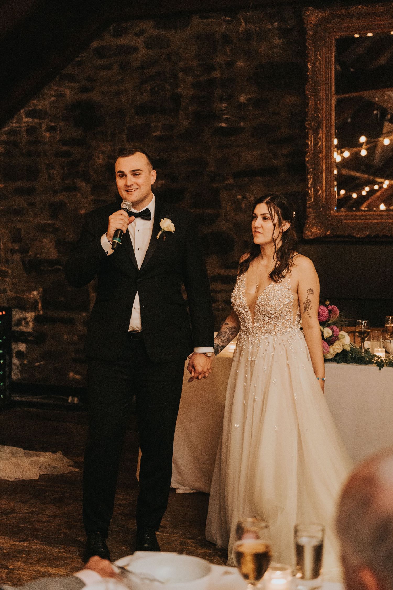Couple in formal wedding attire addresses guests during reception in rustic brick venue.