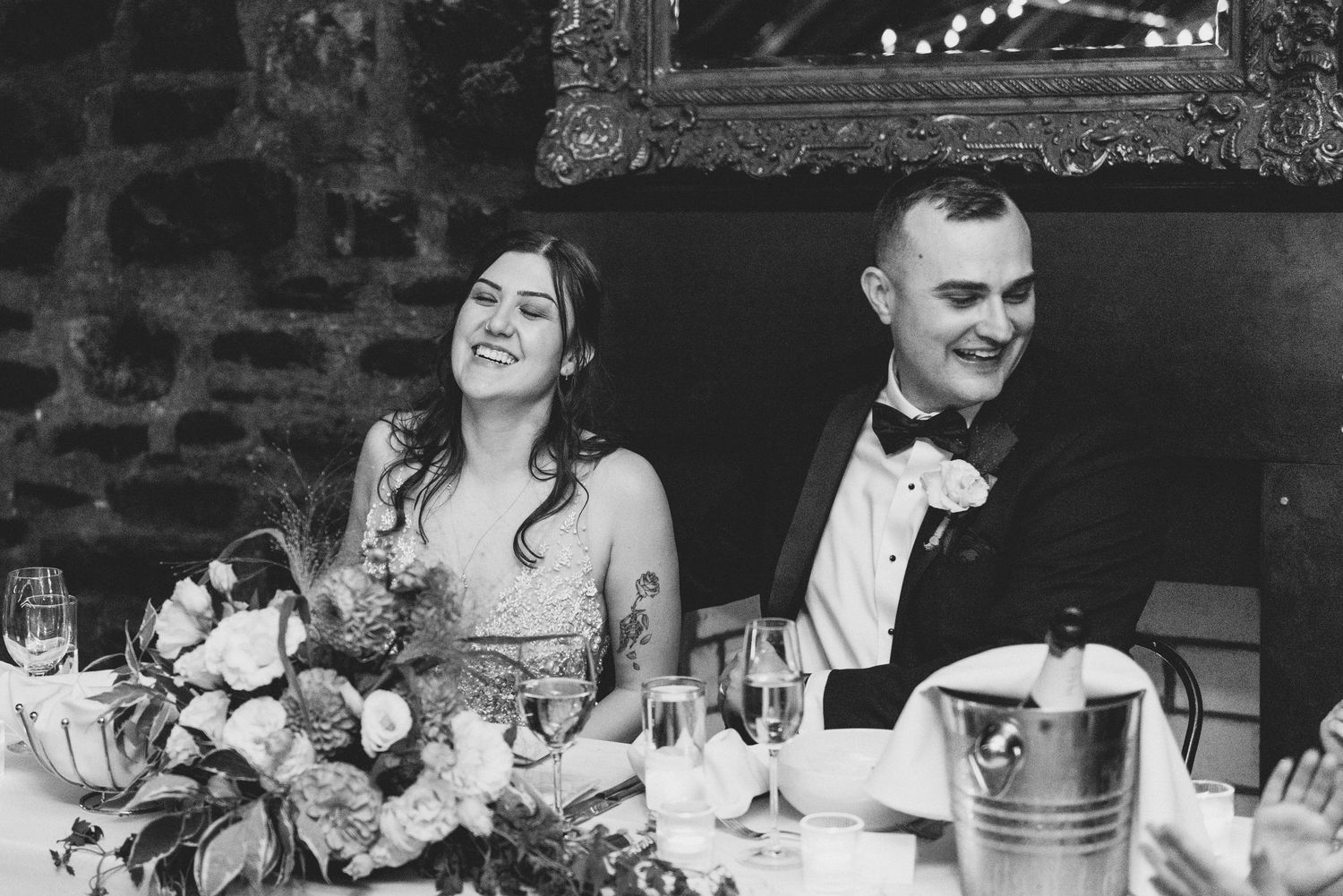A black and white wedding reception photo showing a couple seated at a table with flowers and wine glasses.
