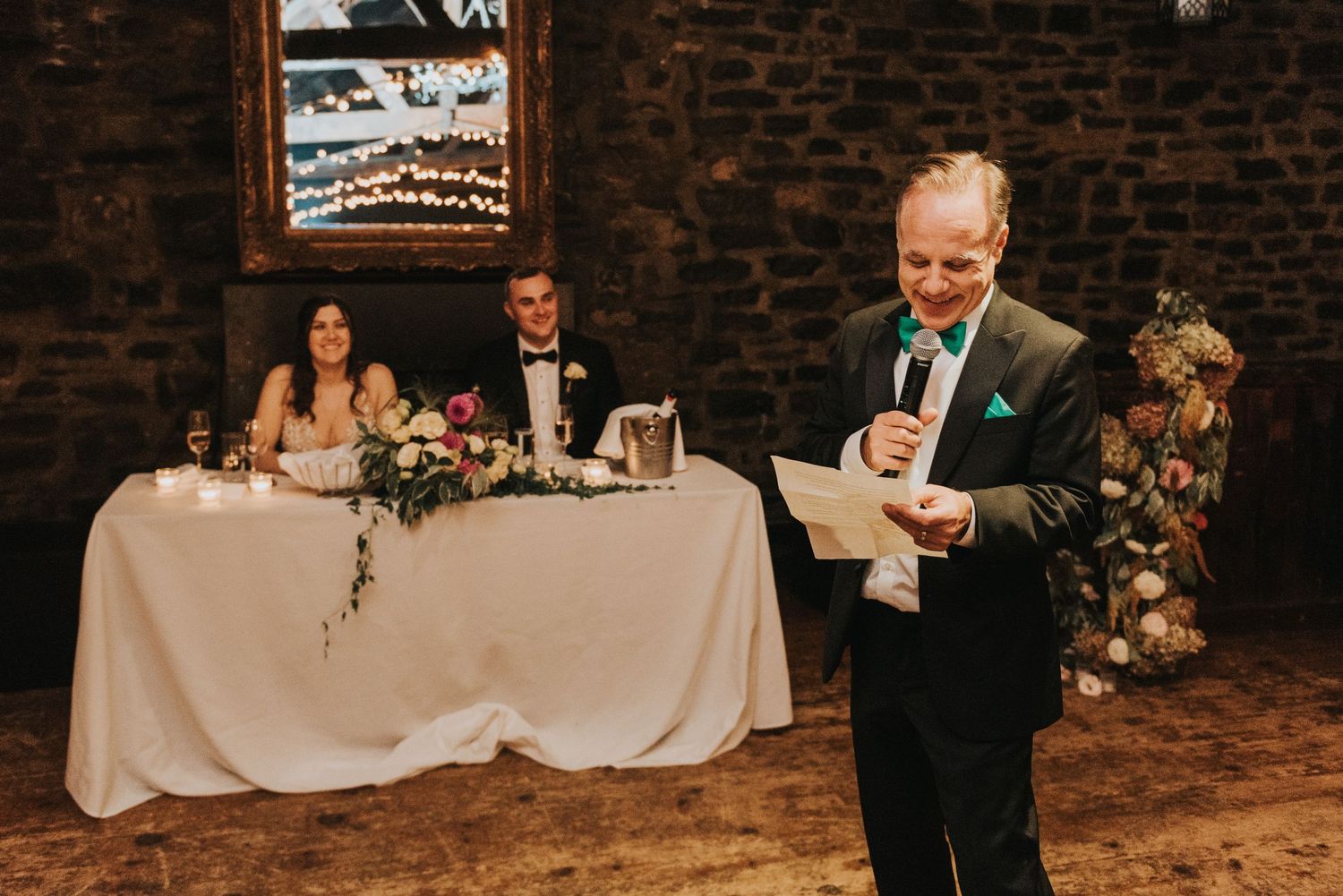 Wedding reception speech being delivered at rustic brick venue with head table and floral decor.
