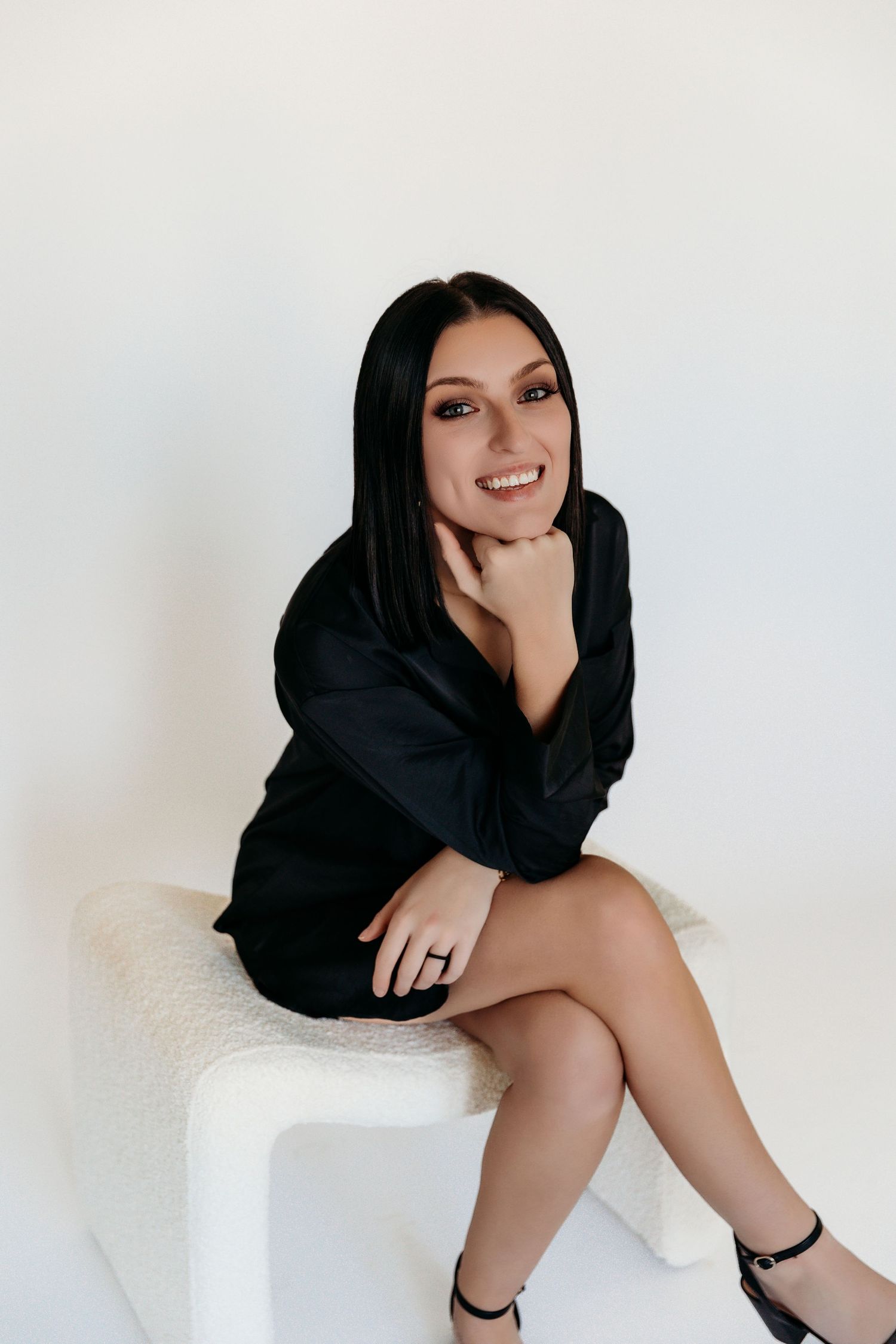 A person in a black dress and dark hair poses against a white background while sitting on a light-colored surface.