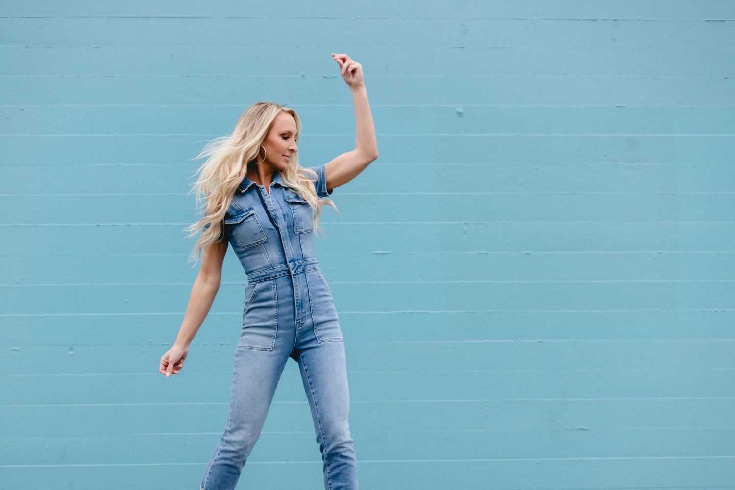 Woman in denim jumpsuit dancing and posing with joy against a bright blue wall.