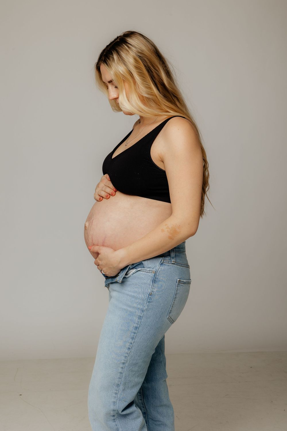 A pregnant woman in blue jeans and a black crop top touches her bare belly against a neutral backdrop.