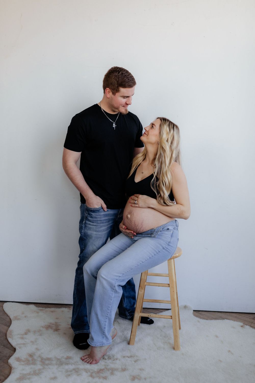 An expectant couple shares an intimate moment as the pregnant woman sits on a wooden stool against a gray wall.