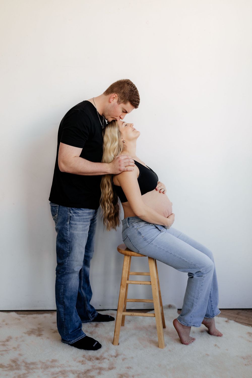 A couple shares an intimate moment as they pose for a pregnancy photo with the woman sitting on a wooden stool.