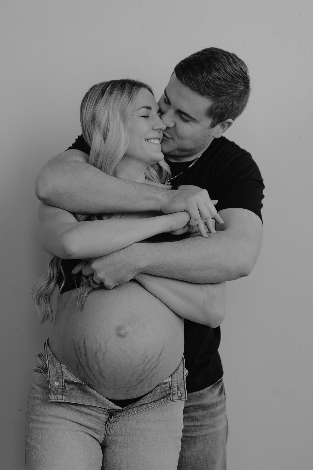 Black and white maternity photo of an intimate embrace showing an exposed pregnant belly in denim.