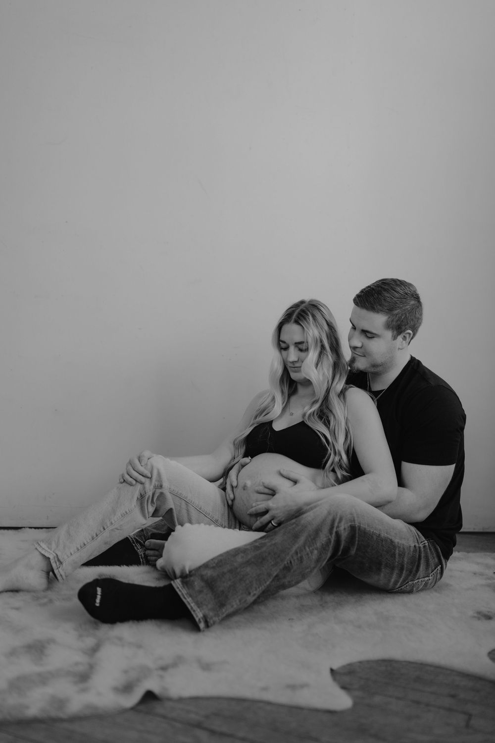 A tender black and white maternity photo of a couple sitting together on the floor in a minimalist setting.