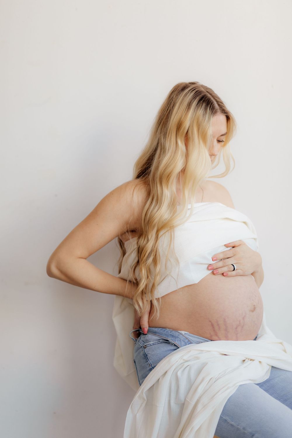 An artistic maternity photo showing an expectant mother in white fabric and jeans against a light background.