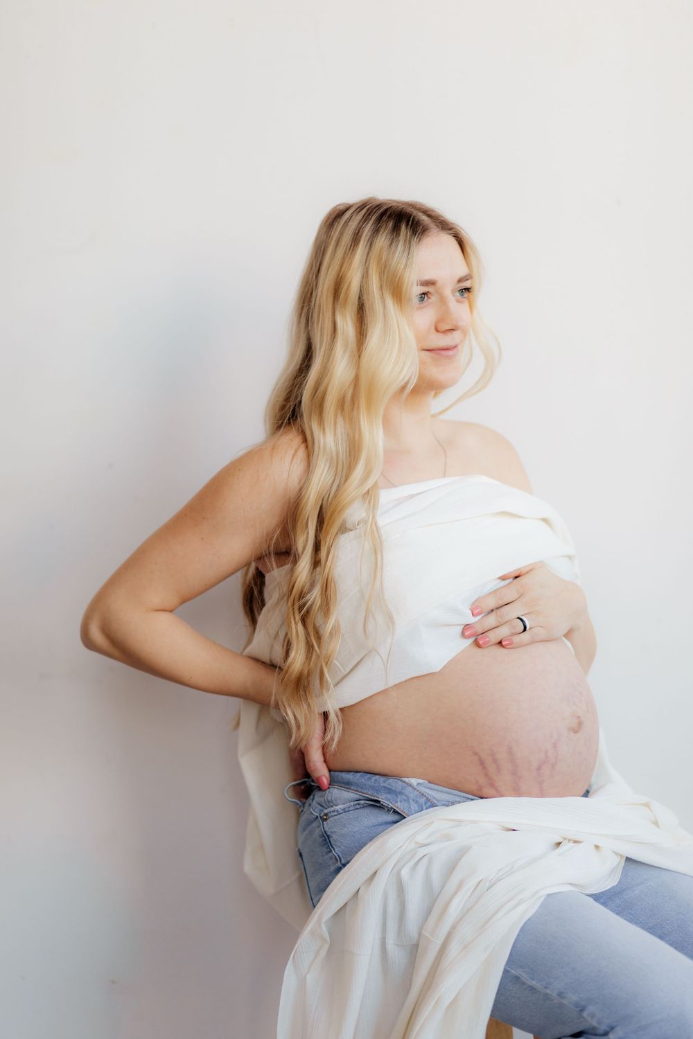 A pregnant woman in jeans and a white shirt poses against a light wall while cradling her belly.