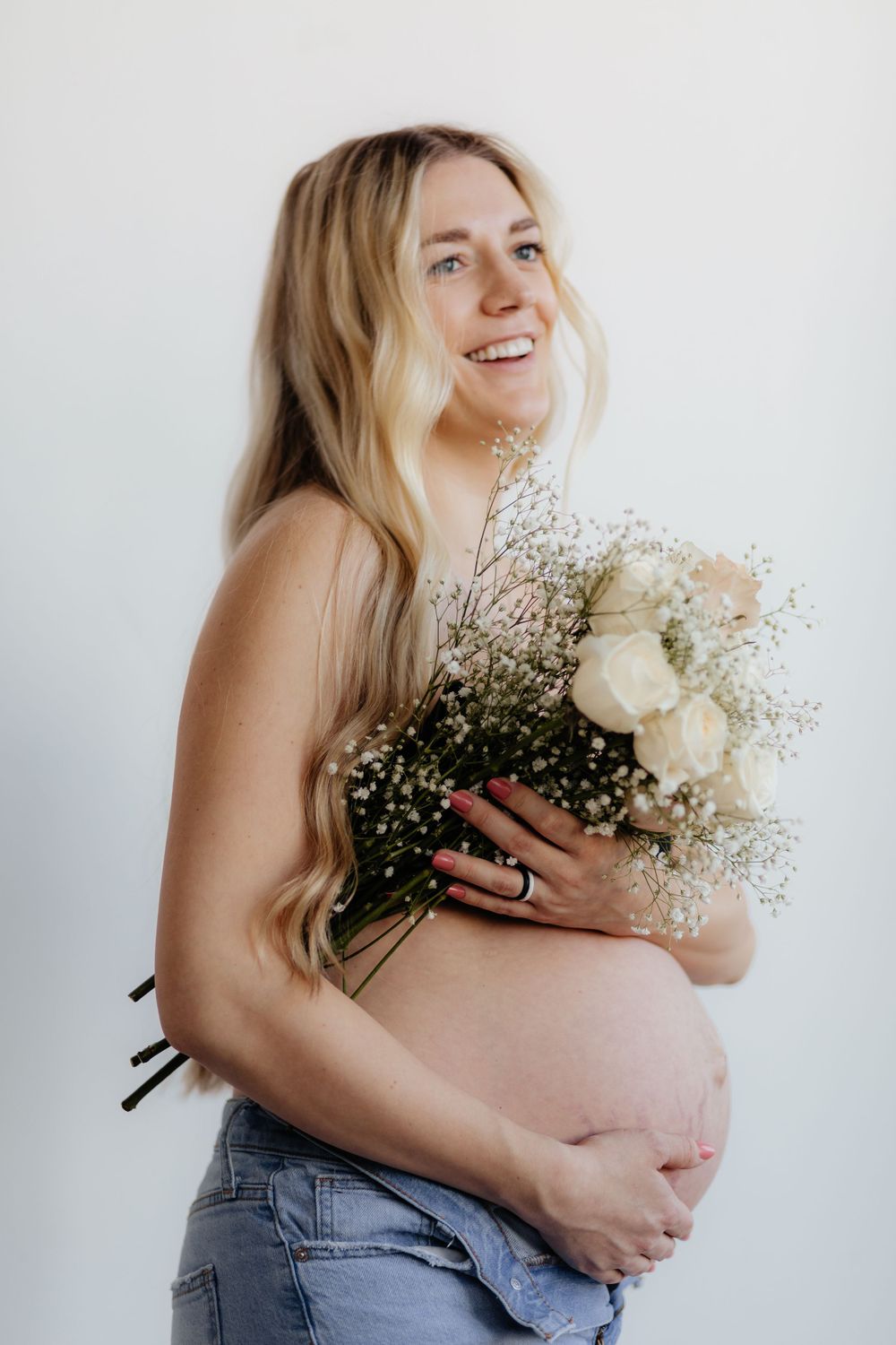 A pregnant woman in jeans holds a beautiful bouquet of white roses and baby's breath against a white background.