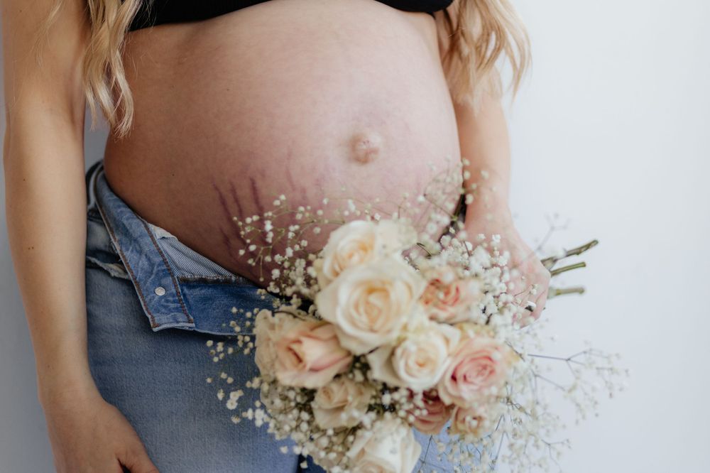 Close-up of pregnant belly with soft peach and cream roses and baby's breath bouquet against denim.
