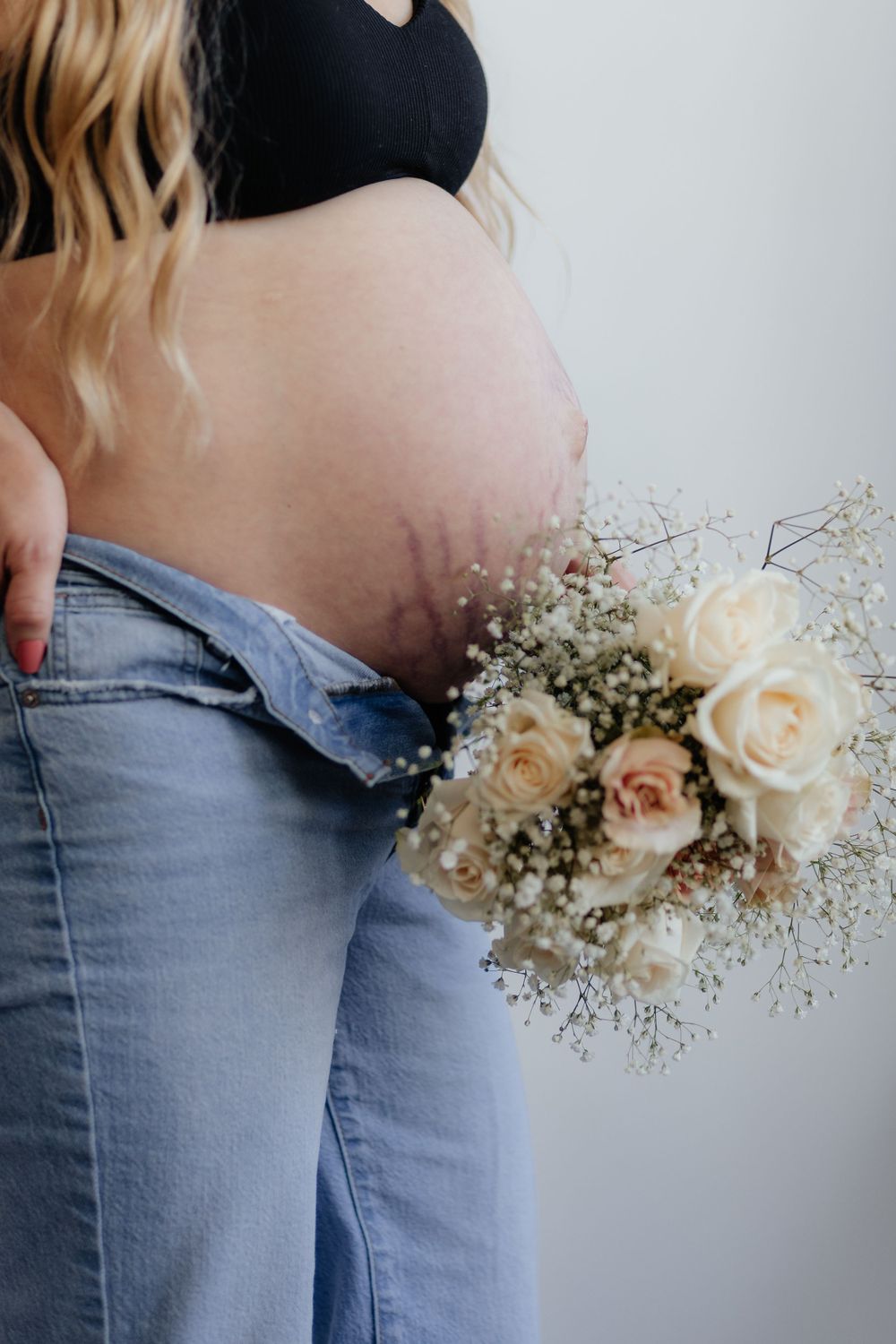 A pregnant woman in jeans holds a delicate bouquet of white roses and baby's breath against her belly.