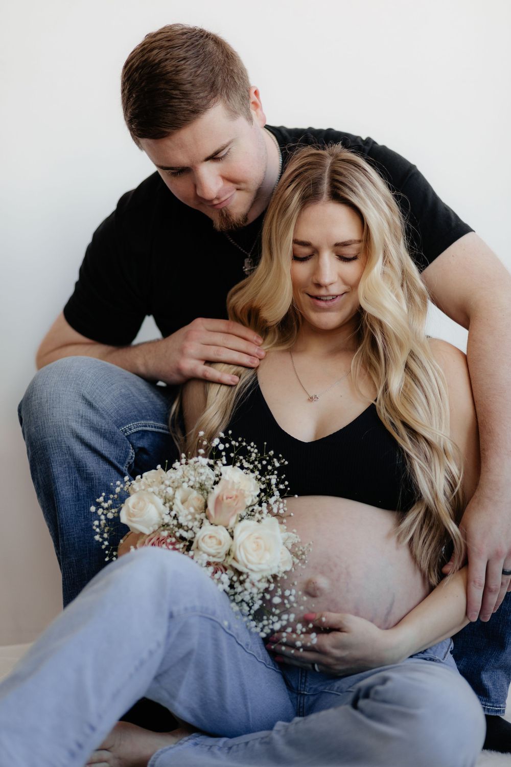 Intimate maternity photoshoot with couple sitting close together holding delicate white roses and baby's breath.