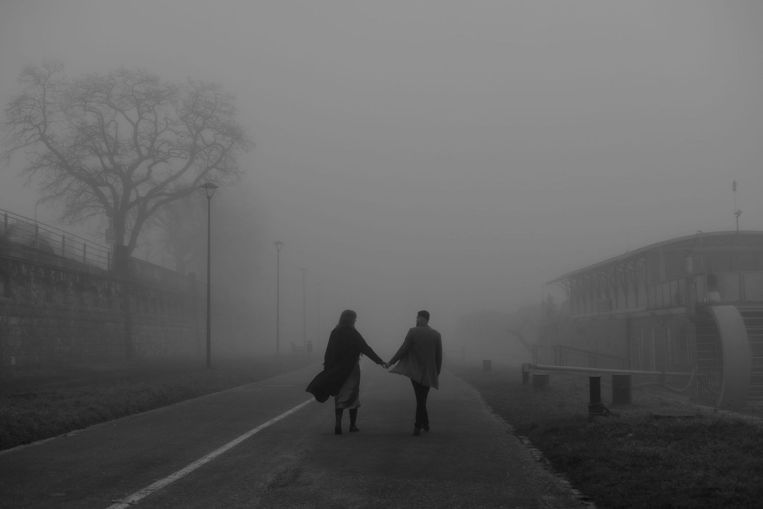Two silhouetted figures hold hands while walking down a misty road on a foggy grayscale day.