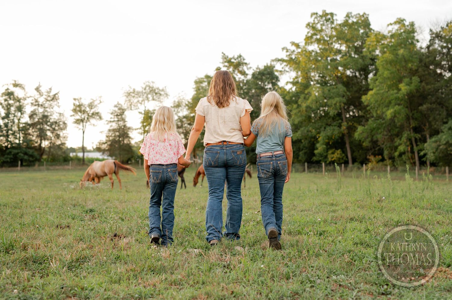 Three people in matching jeans walk away from camera in grassy field with horses visible in background.