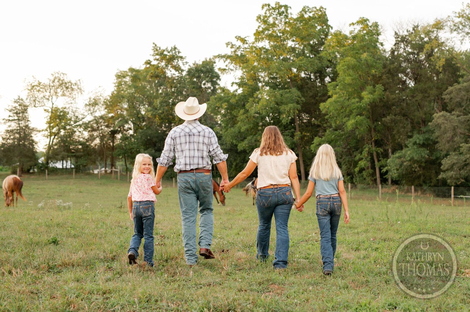 Family walking hand in hand through a pastoral field with horses grazing in the distance.