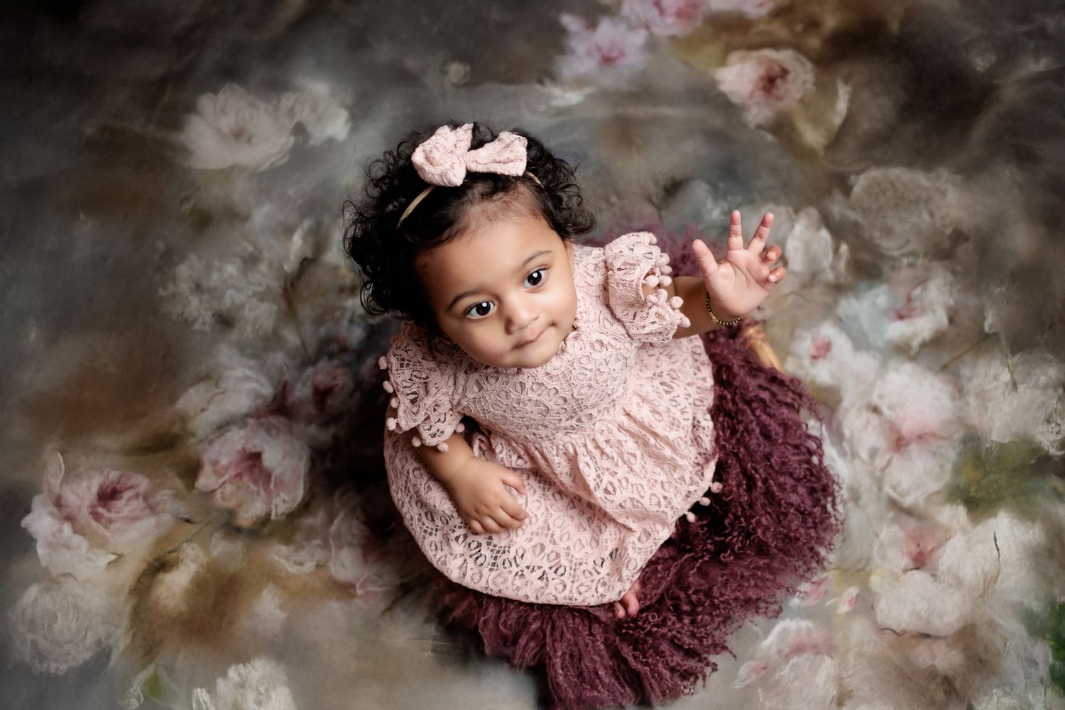 Baby in pink lace dress and burgundy tutu skirt lying on floral backdrop during professional photoshoot.