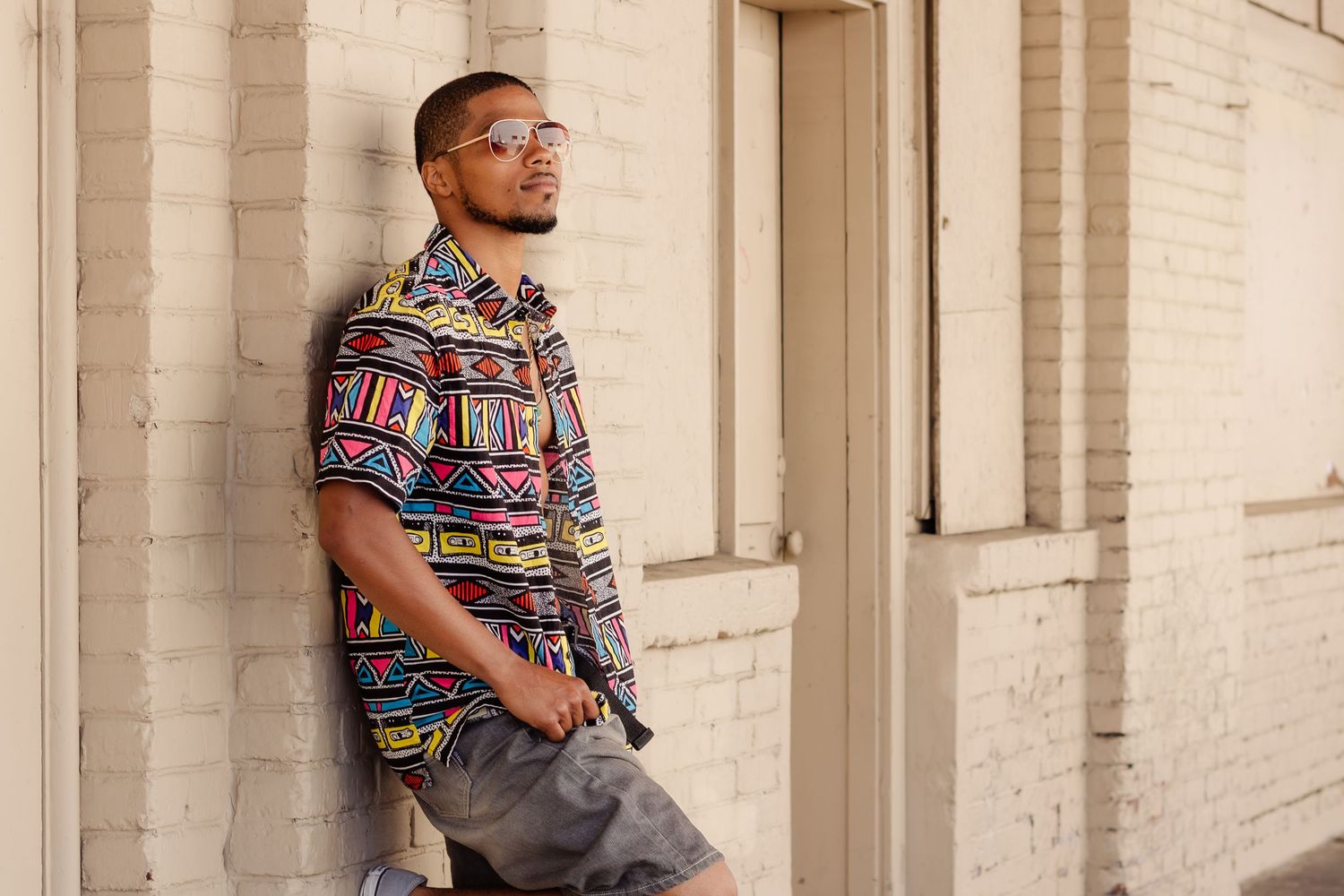Person wearing a vibrant geometric patterned shirt stands against a light brick wall in an urban setting.