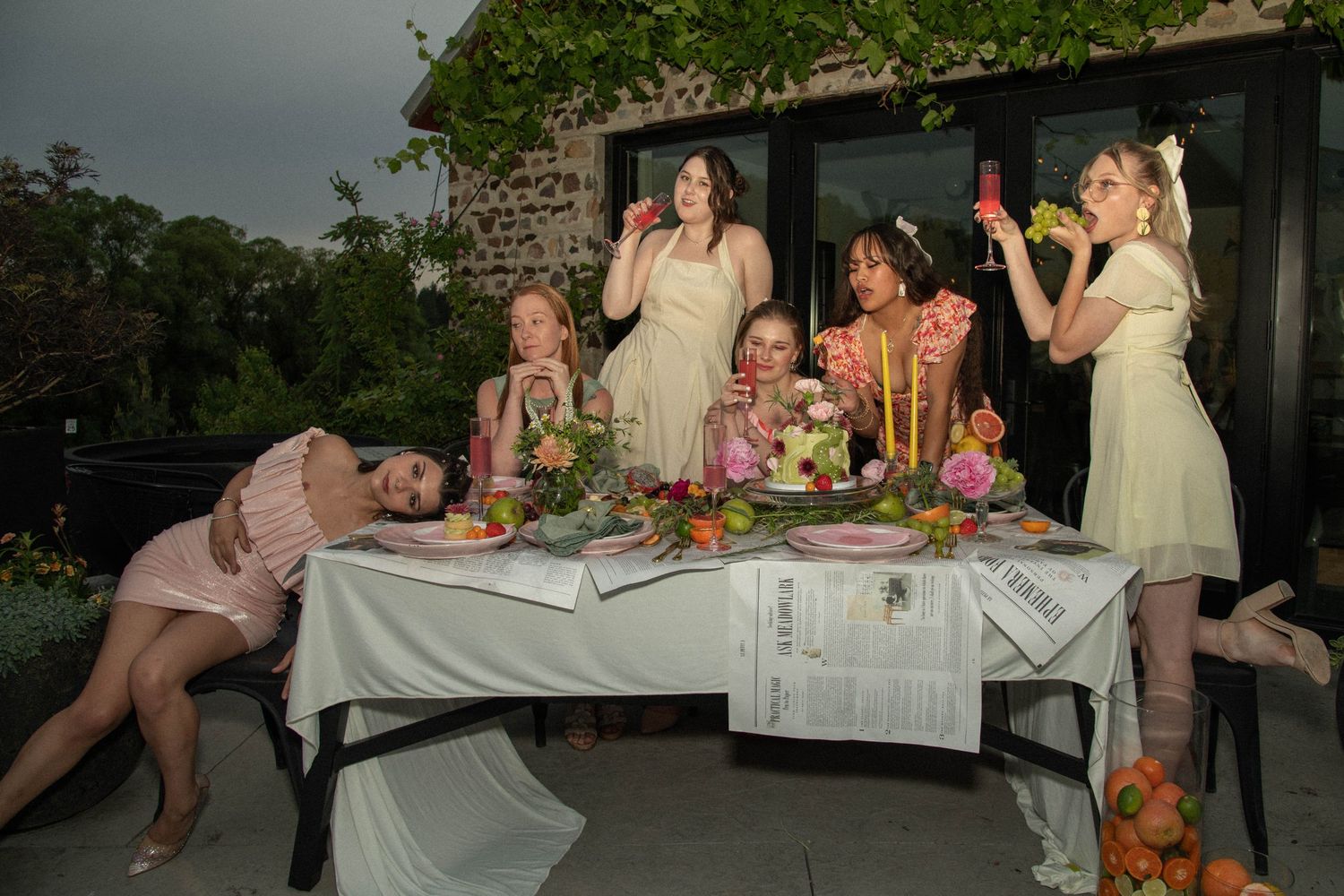 Group of women in pastel dresses gathered around decorated outdoor table with flowers and fruit.