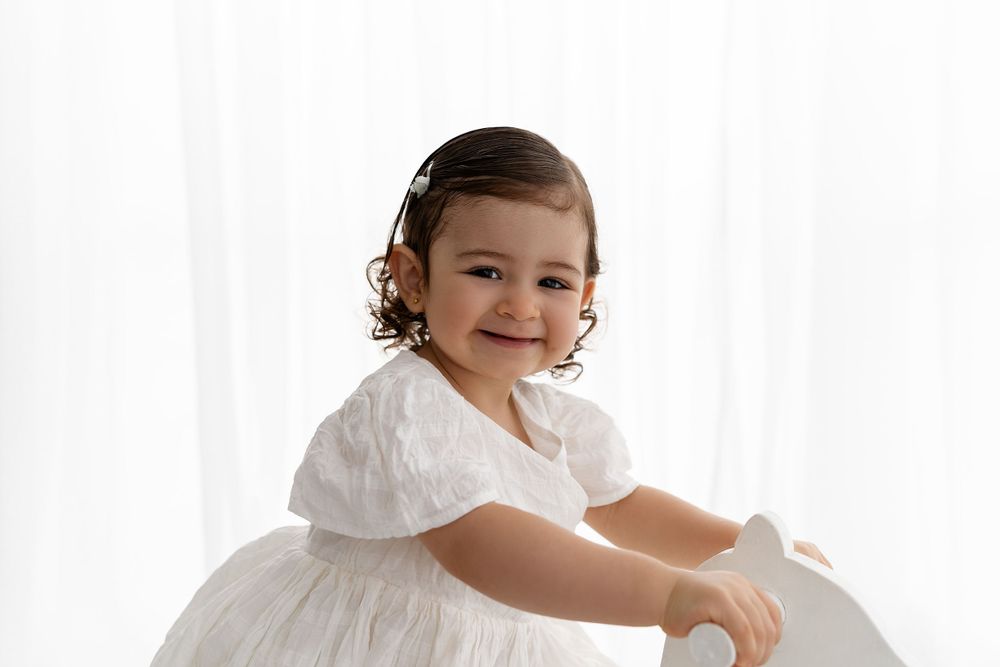 A young child in a flowing white dress sits and smiles against a white background.