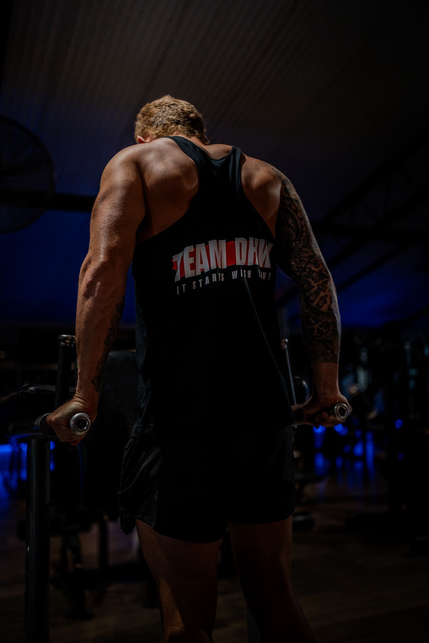 Muscular athlete in Team Dark tank top performs dips exercise at gym with dramatic lighting and blue background.