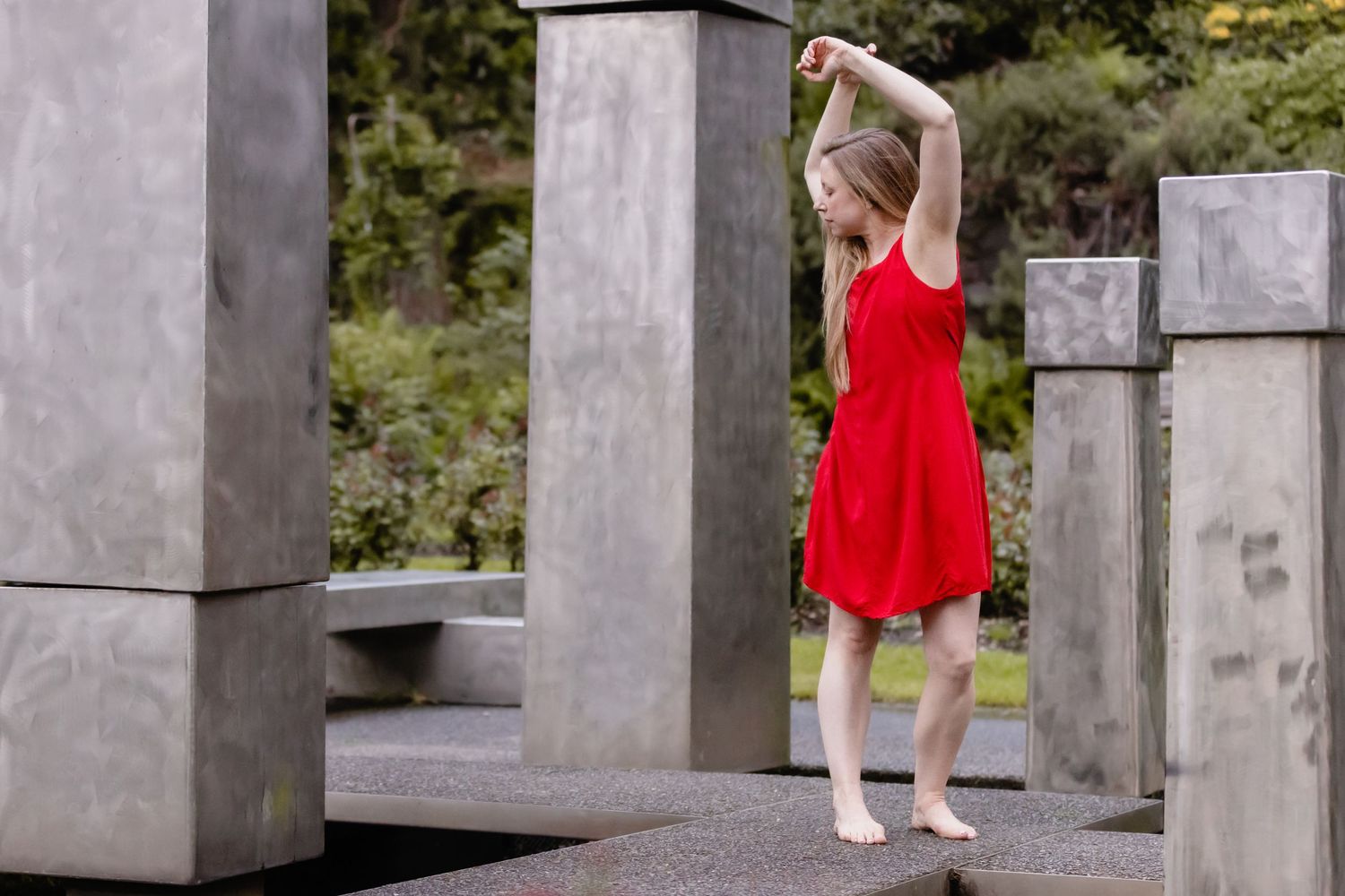 A graceful figure in a flowing red dress dances among modern concrete pillars in an outdoor setting.