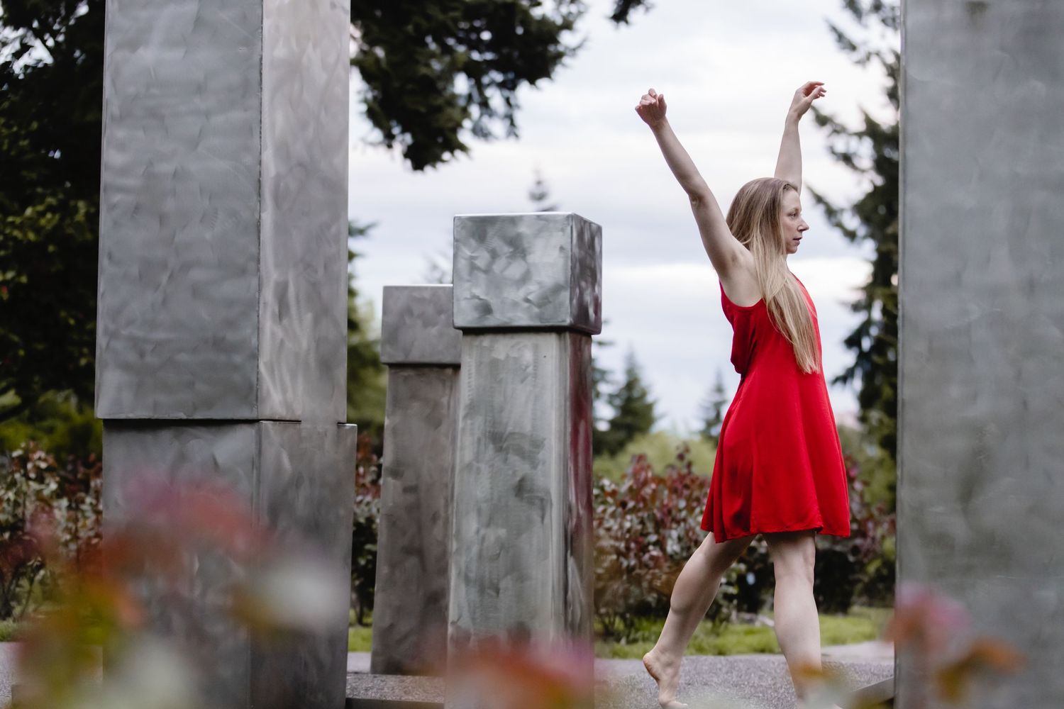 A dancer in a bright red dress performs gracefully among concrete pillars in an outdoor garden setting.