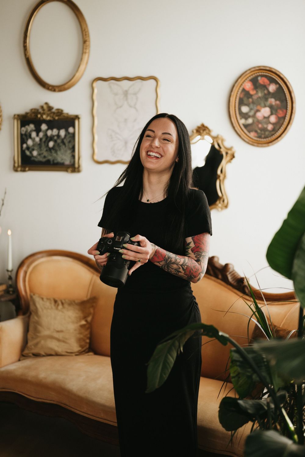 Smiling woman with tattoos in black dress holding camera, standing in elegant room with gold-framed artwork and vintage sofa.
