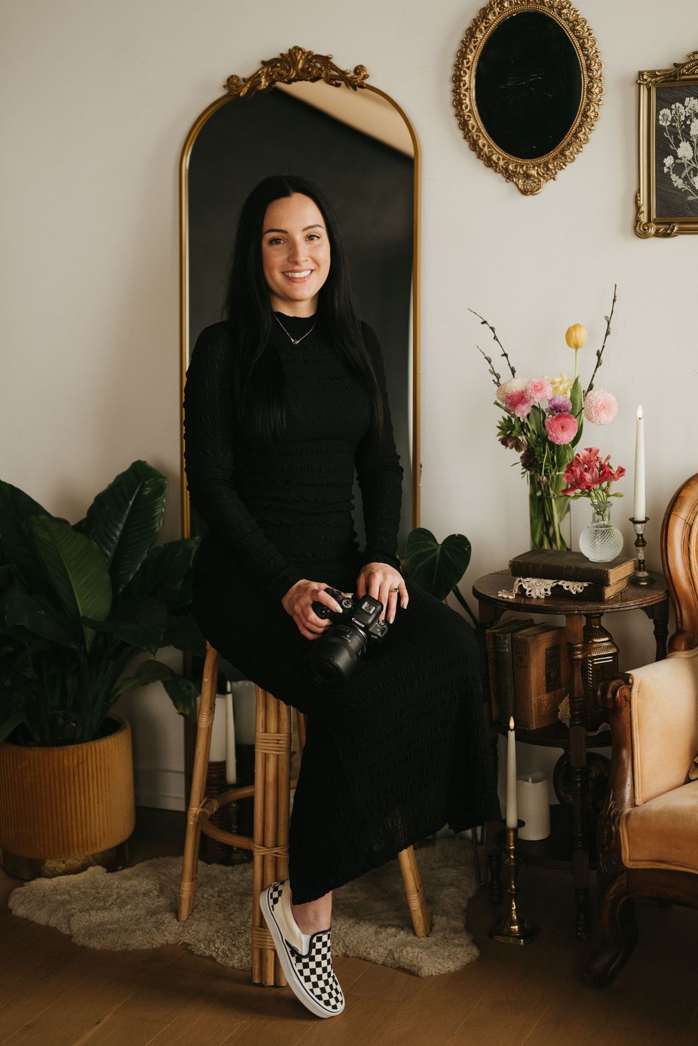 Woman in black dress holding camera sits on stool before gold mirror with flowers and plants nearby.