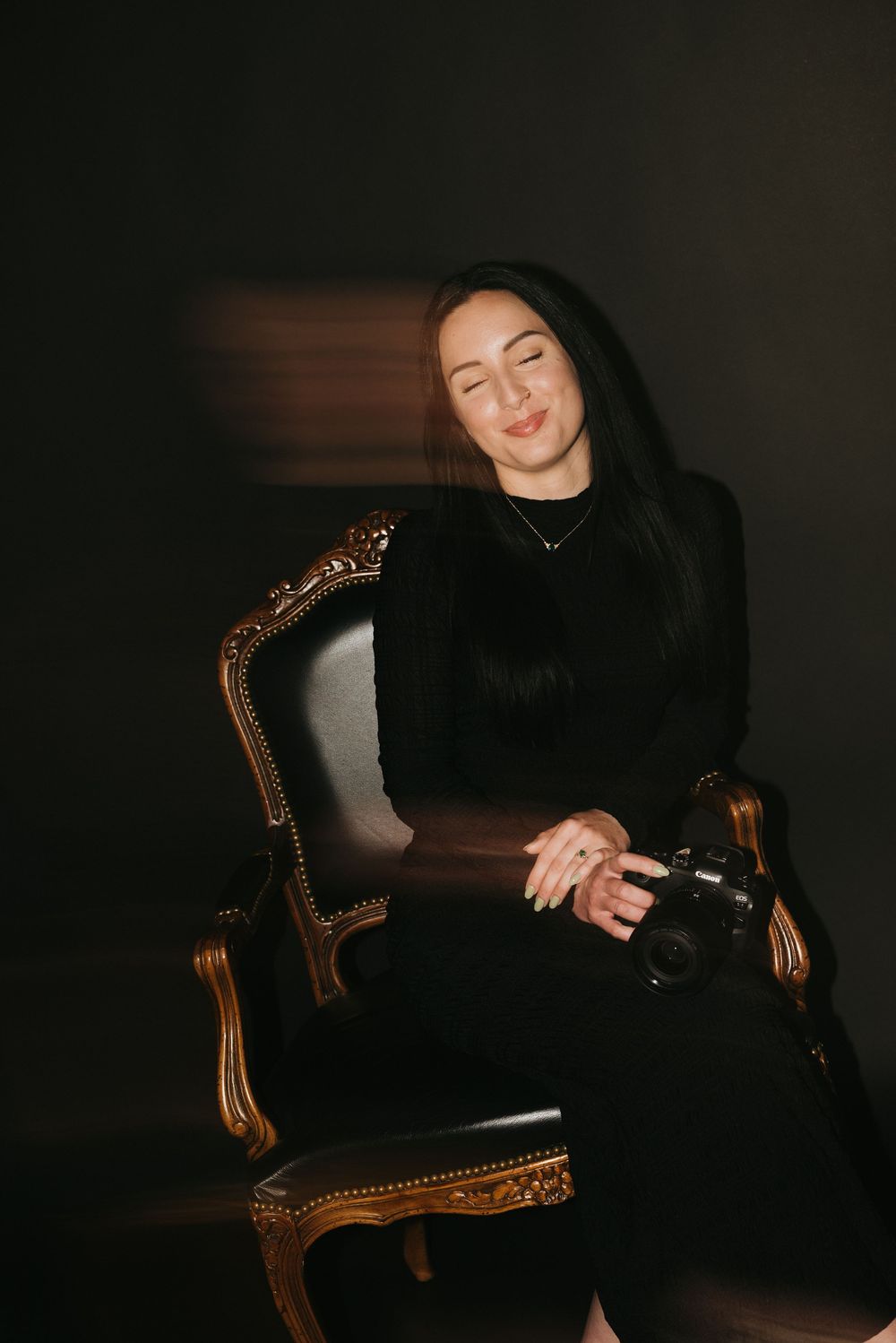 Woman in black dress sits in ornate gold chair holding a camera, illuminated by dramatic studio lighting.