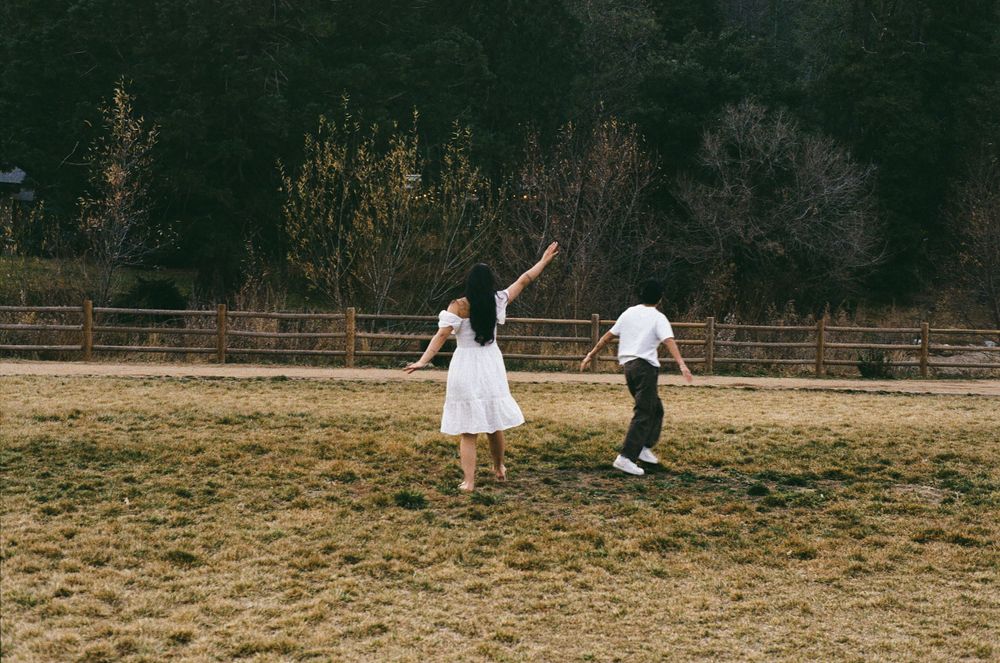Two people dance together in a white dress and dark clothing on a rustic fenced field at dusk.